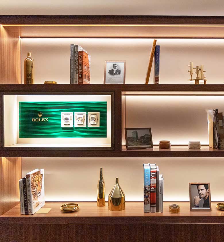 A well-lit wooden bookshelf featuring a mix of decorative items and books. Highlights include a framed green Rolex display, vases, candles, and framed photos. The shelves contain a variety of books, gold decorations, and ceramic pieces, creating an elegant display.