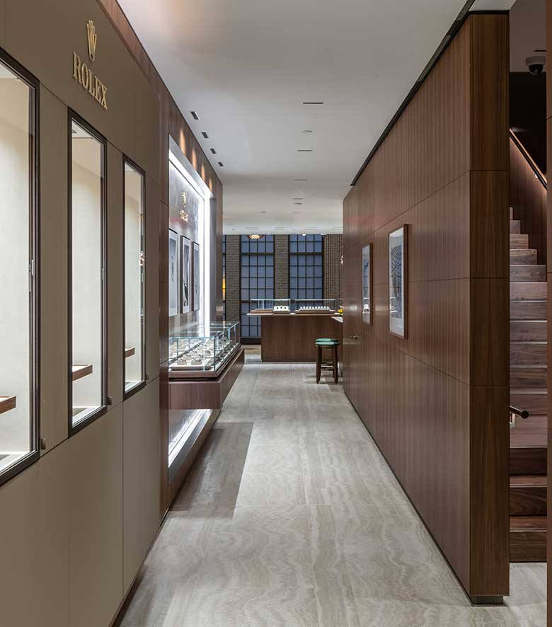 A modern, high-end watch store with wooden walls and flooring. Display cases line the left side, showcasing luxury watches. A counter and bar stool can be seen at the end of the hallway near large windows that let in natural light.