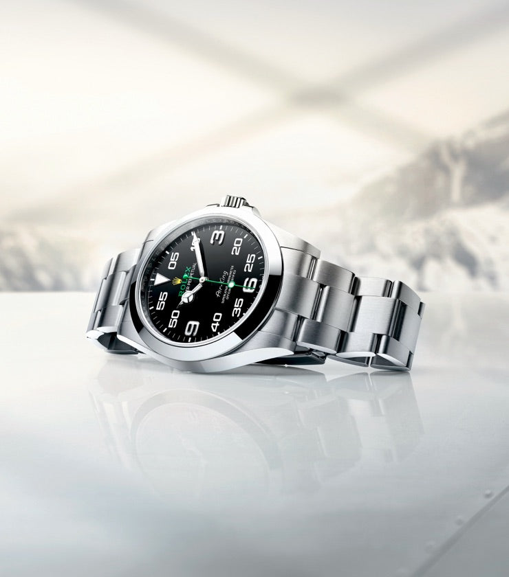 A stainless steel wristwatch with a black dial and white numerals is displayed prominently on a white surface. The background features a blurred mountainous landscape shrouded in snow, evoking a sense of adventure and high altitude.