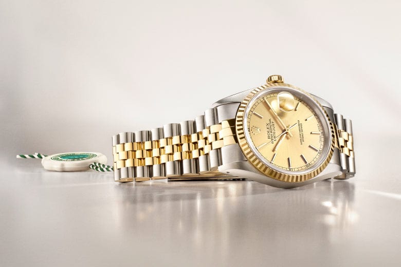 A gold and silver wristwatch with a metal link band is displayed on a reflective surface. A green and white tag is seen in the background.