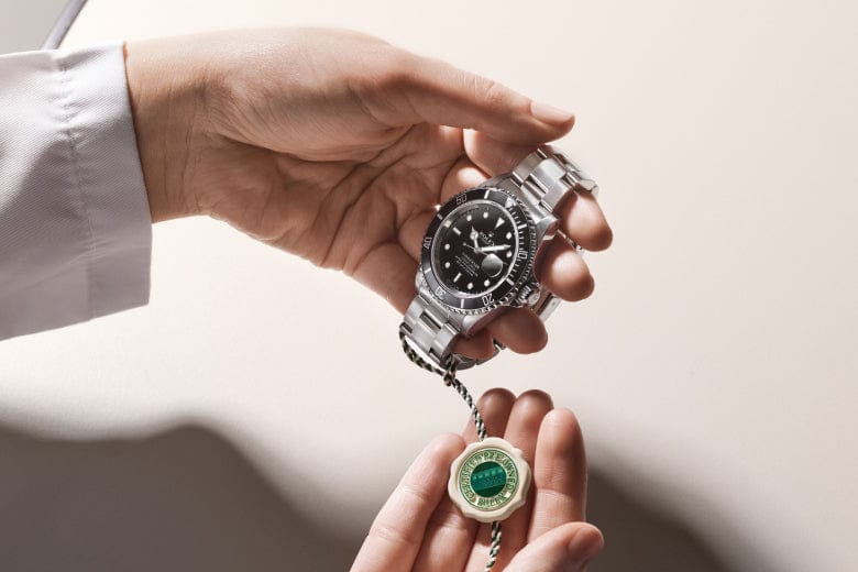 Person holding a silver wristwatch with a black dial and bezel, alongside a green and white tag.