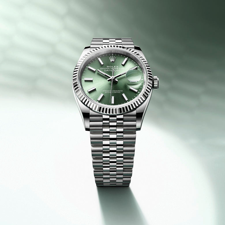 A luxury wristwatch with a stainless steel band and a light green dial is displayed against a green and white gradient background with a textured pattern. The watch features a fluted bezel and a date display window.