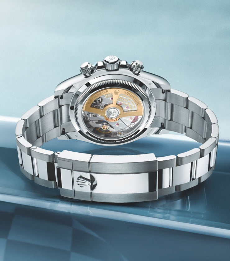 A close-up of a luxury wristwatch with an intricate, exposed mechanical back. The watch has a metallic strap and is placed on a sleek, reflective surface with a gradient background ranging from light to dark blue.