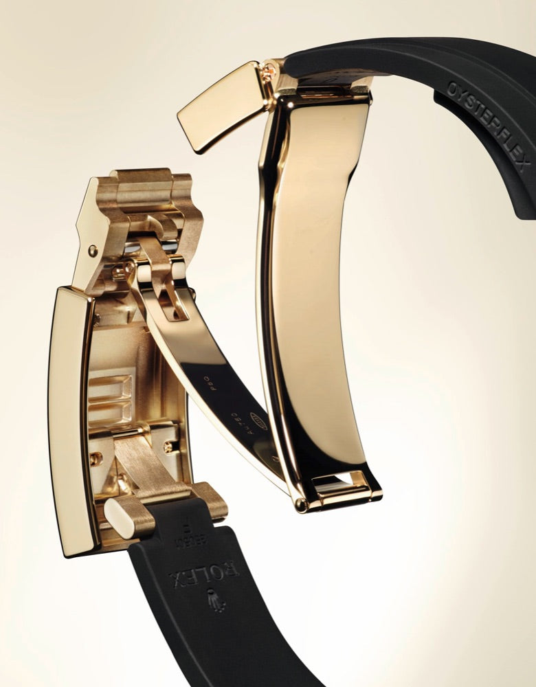 A close-up image of a luxury watch band clasp. The clasp is made of polished gold metal with intricate hinges and a black band. The word 