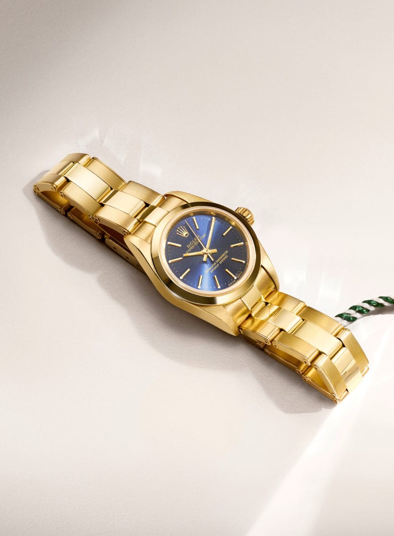 Gold wristwatch with a blue dial and a green tag labeled Certified Pre-Owned Rolex on a light surface.