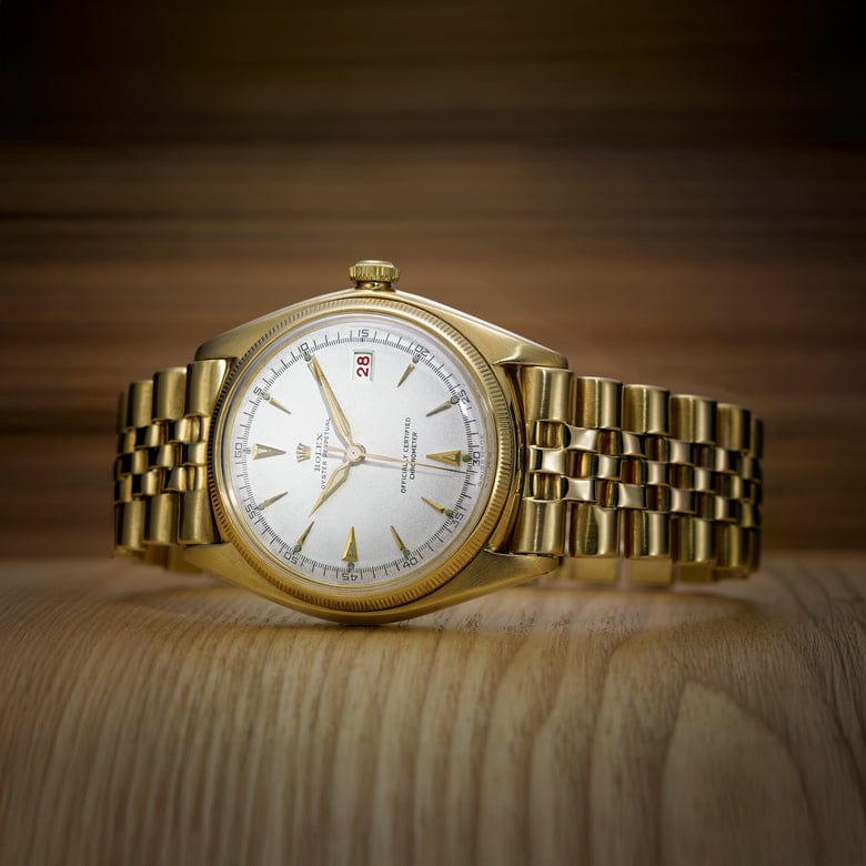 Gold wristwatch with a bracelet-style band, white dial, and Roman numeral hour markers displayed against a blurred wood-textured background.