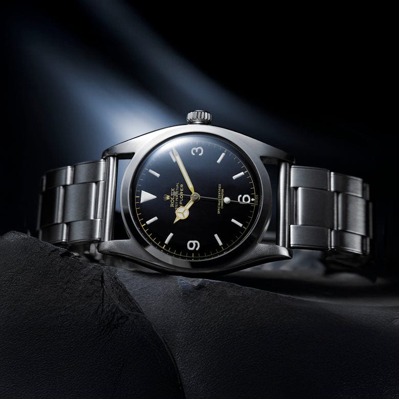 Stainless steel wristwatch with a black dial and luminous markers, displayed on a dark textured surface under focused lighting.