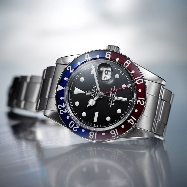 Stainless steel watch with a black dial and a red and blue bezel, displaying the time and date, on a reflective surface.