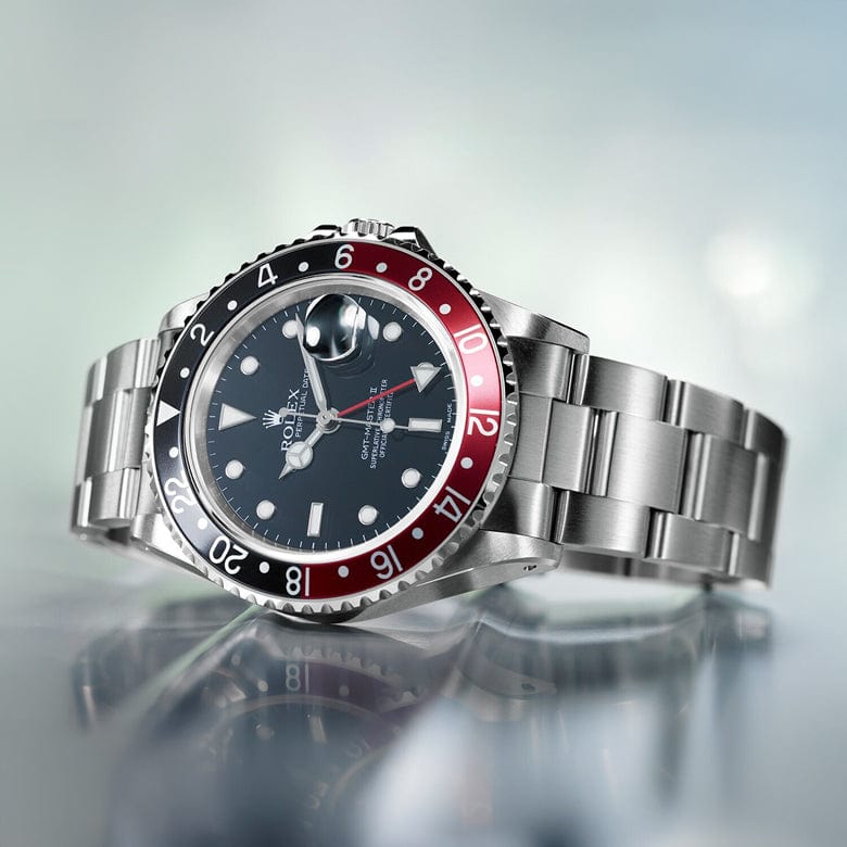 A silver wristwatch with a red and black bezel and a black dial displaying multiple time markers rests on a reflective surface against a blurred background.