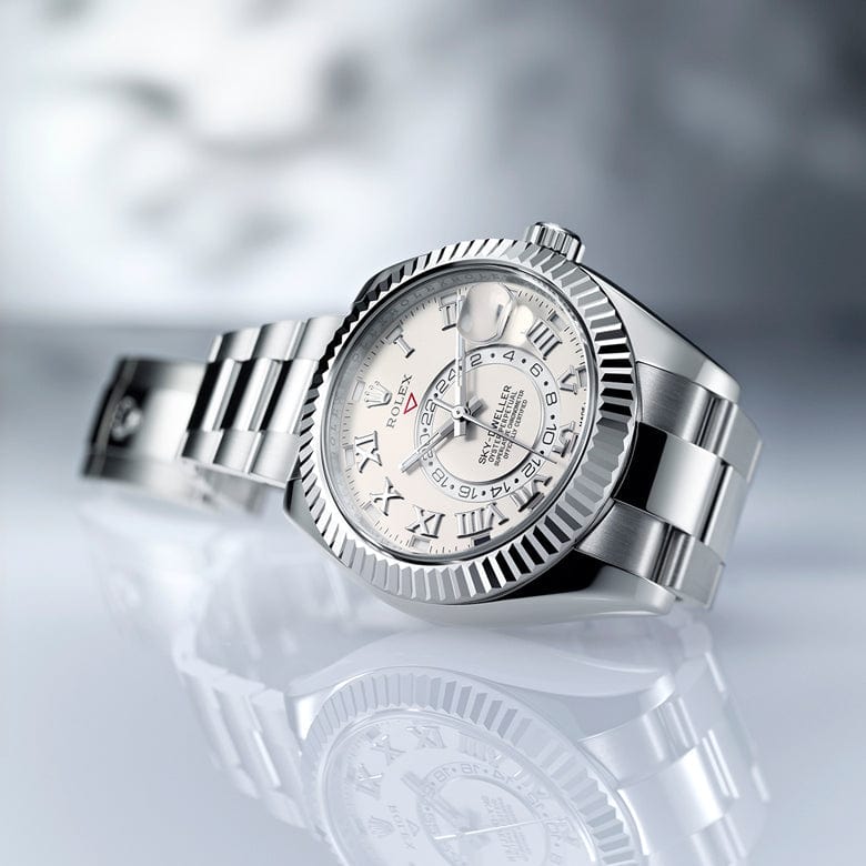 Silver metallic wristwatch with a textured bezel, white dial, Roman numerals, and an ornate central pattern, resting on a reflective surface.