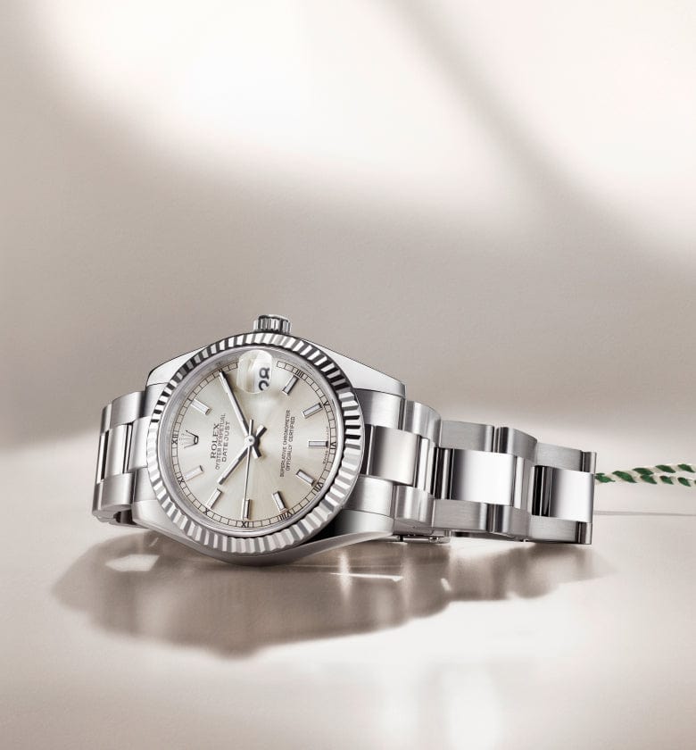 A silver wristwatch with a white dial and metallic band lies on a reflective surface, next to a green and white tag.