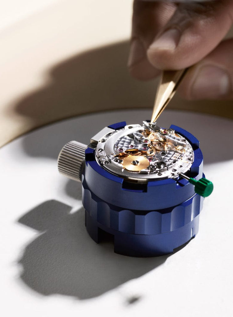 A hand uses tweezers to adjust a mechanical watch movement on a blue holder.