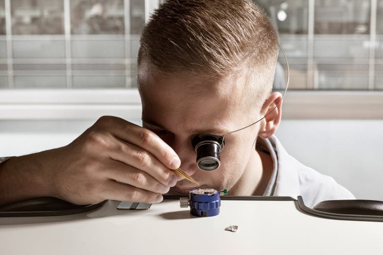 A person uses a magnifying eyepiece to carefully work on assembling a small mechanical device on a table.