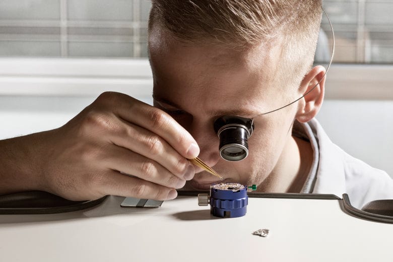 Person using a magnifying lens and tweezers to work on a small mechanical component, possibly a watch or clock part, on a table.