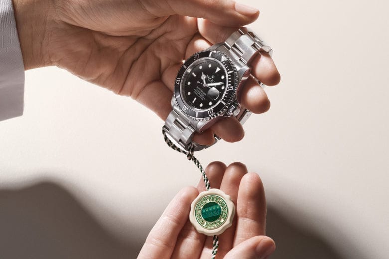 Hands hold a luxury watch with a stainless steel band and a black dial. A green and white tag is attached to the watch.