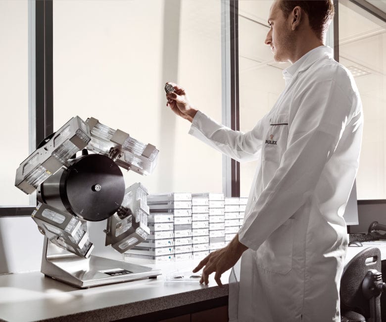 A person in a lab coat inspects a small round object while standing beside advanced machinery and stacks of silver rectangular items on a countertop in a well-lit room.