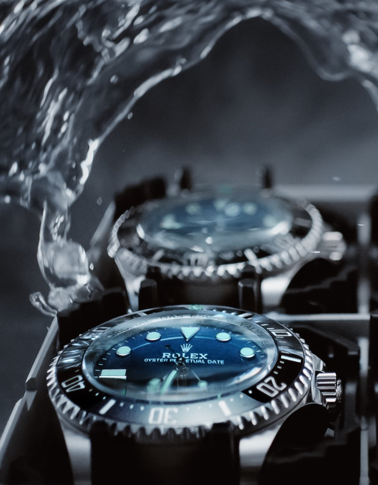 A close-up shot of multiple luxury watches with blue dials arranged in a box, partially submerged and surrounded by splashing water, emphasizing their water-resistant feature. The watches have metallic bands and bezel markings with prominent hour markers.