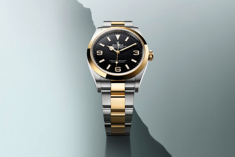 A luxury wristwatch with a black dial and gold numerals, encased in a polished silver and gold two-tone metal band, is placed against a gradient grey background. The watch displays the current time and the brand‚Äôs logo is visible on the dial.