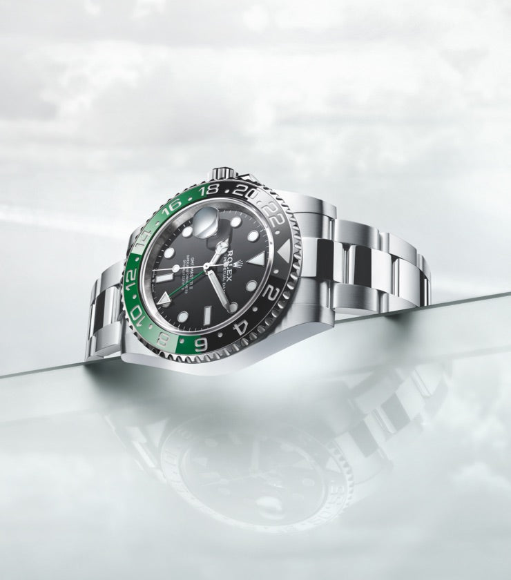 A luxury stainless steel wristwatch with a black dial and green and black bezel, featuring luminescent hour markers and hands. The watch is elegantly displayed on a sleek glass surface against a light, cloudy background.