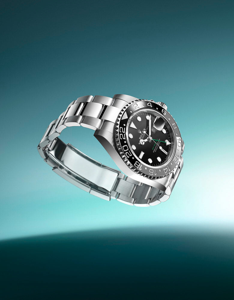 A luxury stainless steel wristwatch with a black dial and bezel featuring luminous markers. The watch is angled artistically against a gradient blue-green background, giving it a floating appearance. The bracelet is metal with polished and brushed finishes.