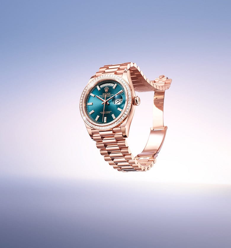 A luxurious, rose gold wristwatch with a link bracelet and a teal face is suspended against a gradient light blue background. The watch features Roman numeral markers and a date window on the right side of the dial.