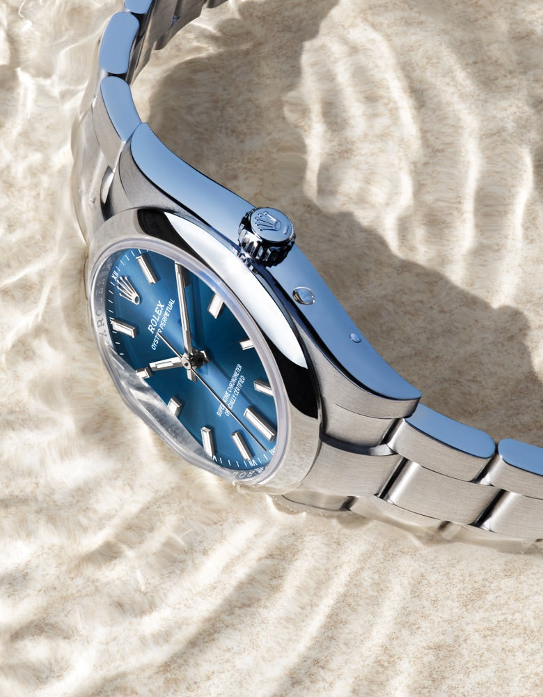 A close-up of a silver wristwatch with a blue dial set against a textured, light-colored background. The watch has a metal band and a classic design with a knurled crown.