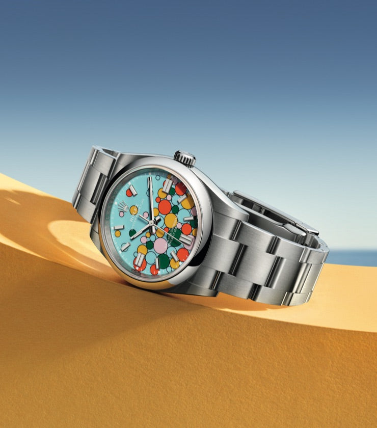 A silver wristwatch with a colorful dial featuring various colorful circular shapes is displayed on a light yellow, curved surface against a blue sky background. The watch has a metal band and a minimalist, modern design.