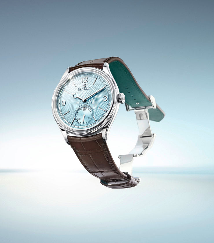 A luxury wristwatch with a light blue dial, intricate detailing, and a brown leather strap is displayed on a blurred blue and gray background. The watch features metallic hour markers and hands, with a subdial above the 6 o'clock position.