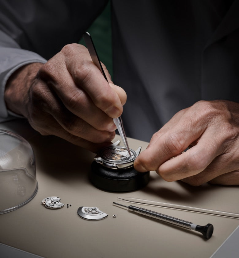 A skilled watchmaker's hands delicately work on the components of a mechanical watch. Precision tools, tiny screws, and watch parts are seen on the table, along with an open watch mechanism, highlighting the intricate craftsmanship involved.