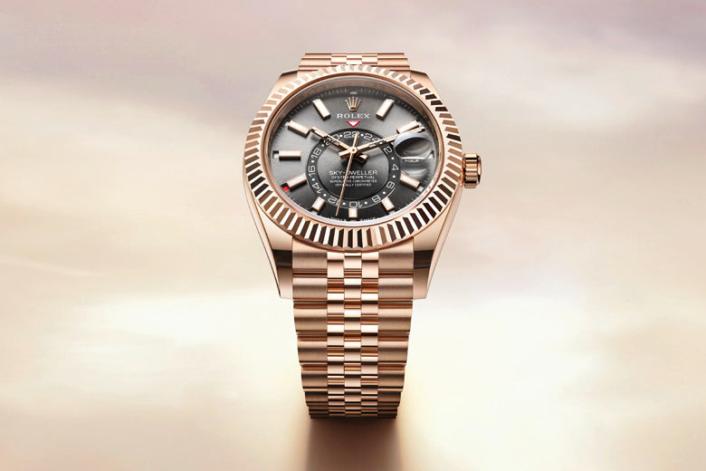 A luxurious gold wristwatch with an intricate gold band and a dark, detailed dial is displayed against a softly blurred, light-colored background. The watch features a prominent gold bezel and appears very high-end and sophisticated.