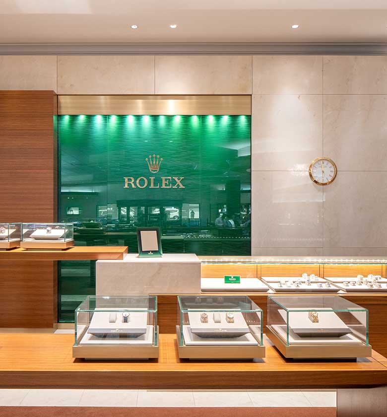 A luxurious store display featuring glass cases with various Rolex watches. The background has a green wall with the Rolex logo, and a clock hangs on a marble wall to the right. The modern interior includes wooden and marble elements.