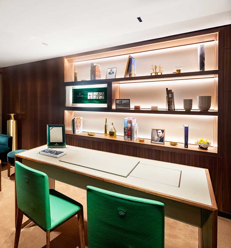 A modern office with a green velvet sofa, matching seating, a wooden coffee table, and a contemporary desk. The background features wooden paneled walls with various decor items, books on shelves, and a lit display case. The space is well-lit and stylish.